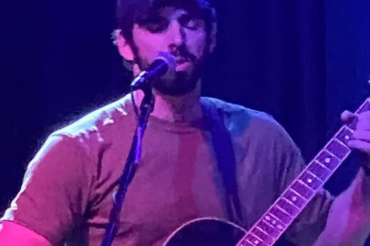 A musician is performing on stage, playing an acoustic guitar and singing into a microphone, with dim lighting creating a moody atmosphere.