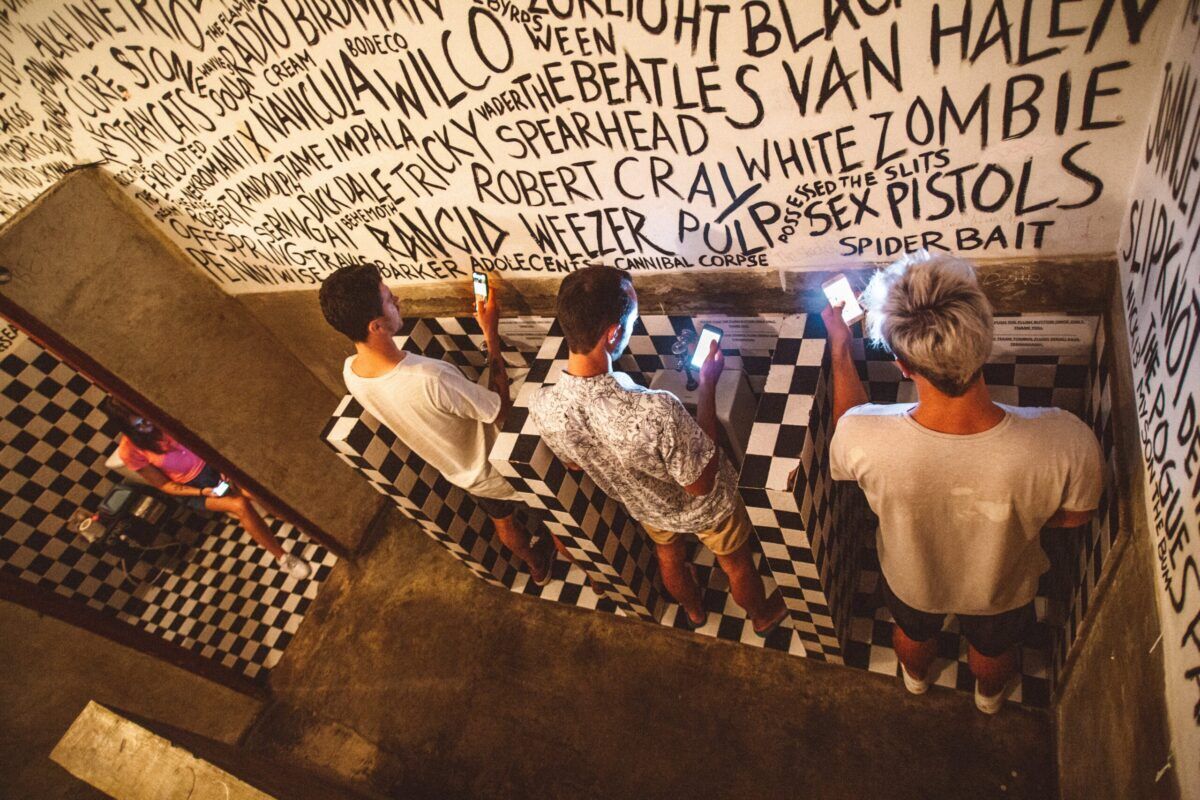 The image shows three people standing in a stairwell, surrounded by black and white checkered flooring and walls covered in music-related graffiti.