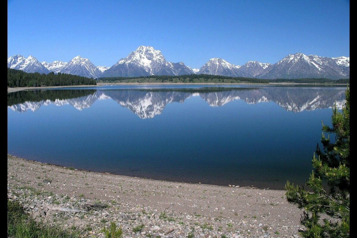 A tranquil lake reflects snow-capped mountains under a clear blue sky, framed by a sandy shore and lush greenery.