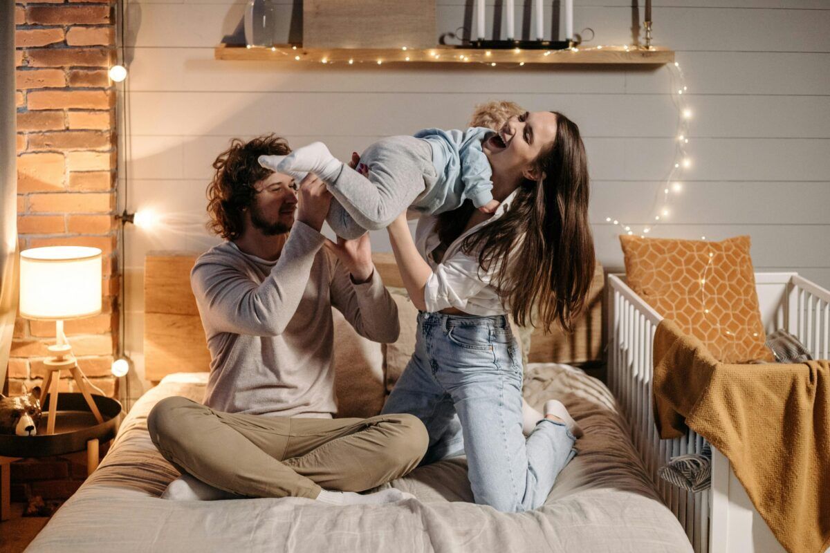 A couple plays joyfully with their child on a bed, surrounded by warm lighting and a cozy atmosphere. The scene captures a moment of family bonding and happiness.