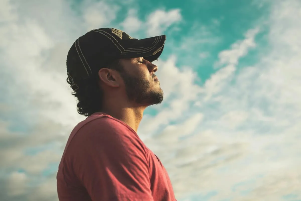 A young man in a cap gazes upward, with clouds and a blue sky in the background, evoking a sense of contemplation and serenity.