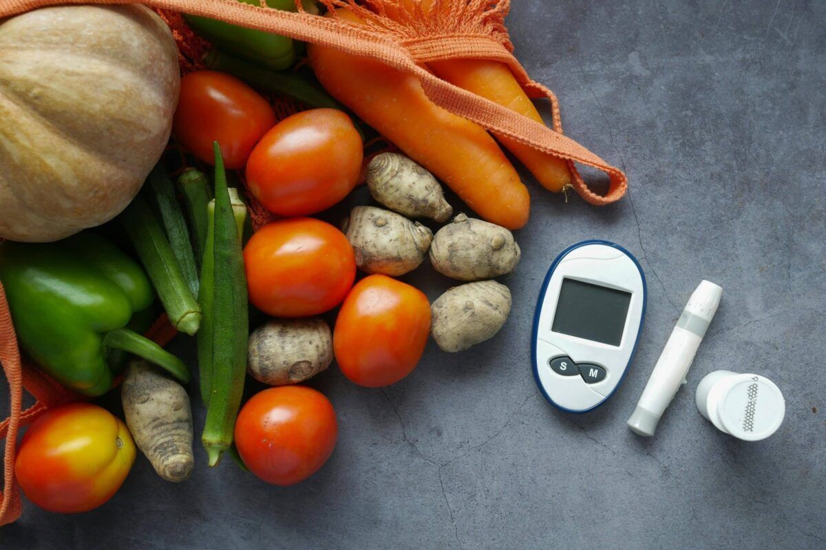 A collection of fresh vegetables, including tomatoes and peppers, accompanied by a blood glucose monitor and diabetes medication on a gray surface.