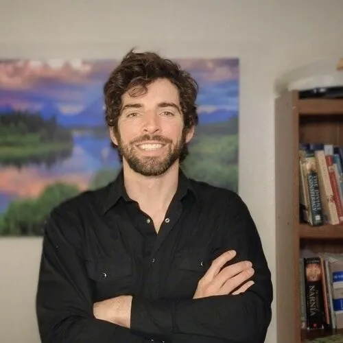 A smiling Colt Gordon with curly hair stands with crossed arms in front of a colorful landscape painting, next to a bookshelf filled with books.